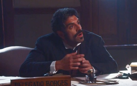 O ator Danilo Dal Farra sentado à uma escrivaninha, com expressão de surpresa, como Borges em cena de Nos Tempos do Imperador