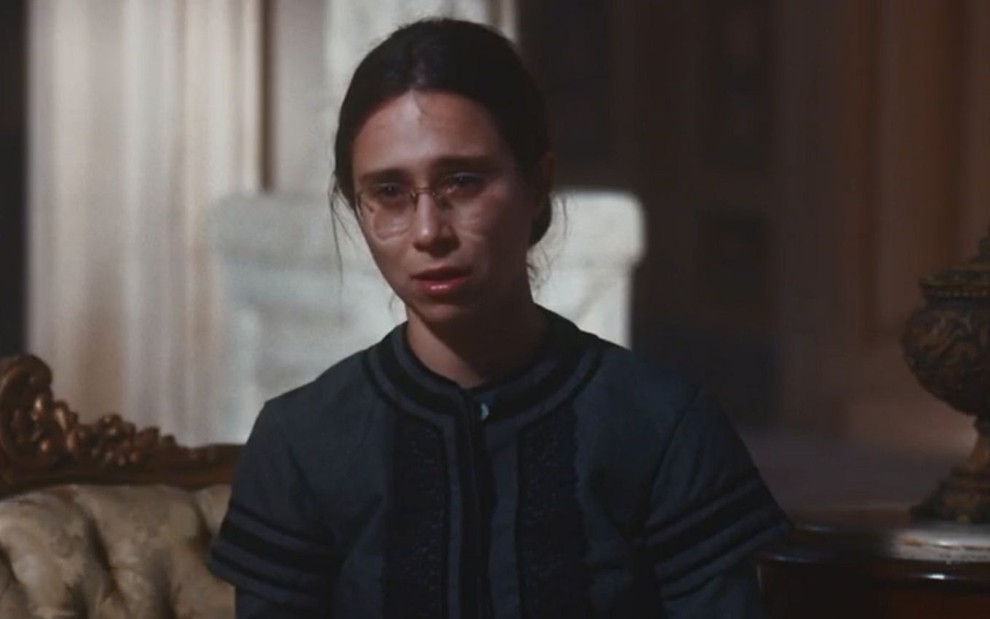 Dolores (Daphne Bozaski) está sentada na sala da Quinta da Boa Vista; ela chora em cena de Nos Tempos do Imperador