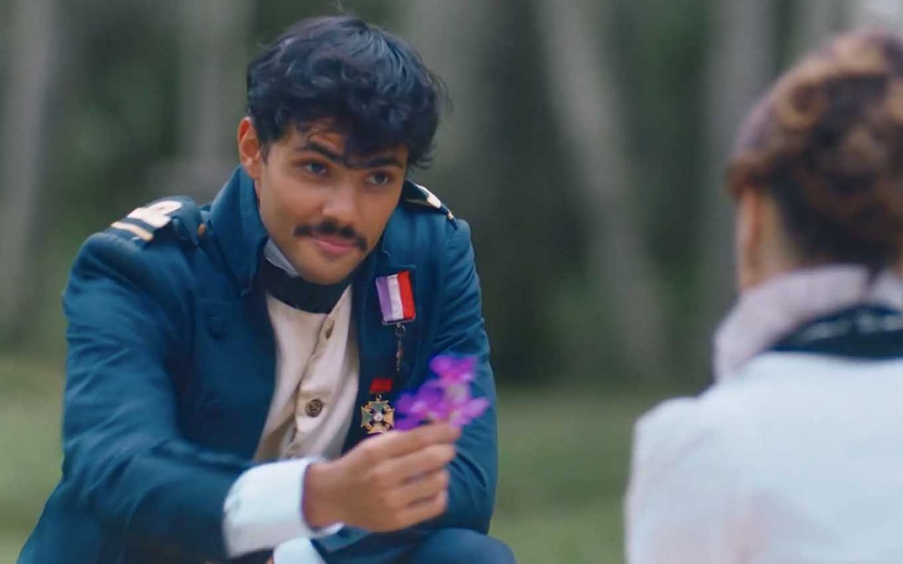 O ator Gabriel Fuentes, com uma farda de marinheiro, entrega uma flor como o Bernardinho para Bruna Griphao, de costas como a Leopoldina, em Nos Tempos do Imperador