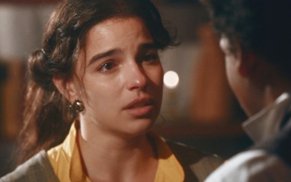 Pilar (Gabriela Medvedovski) está em pé na sala da casa de Samuel (Michel Gomes), ela chora ao conversar com o ex-noivo, que está de costas na imagem