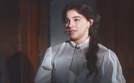 Pilar (Gabriela Medvedovski) está em pé na recepção de hotel de Quinzinho (Augusto Madeira), ela usa uniforme de médica em cena de Nos Tempos do Imperador