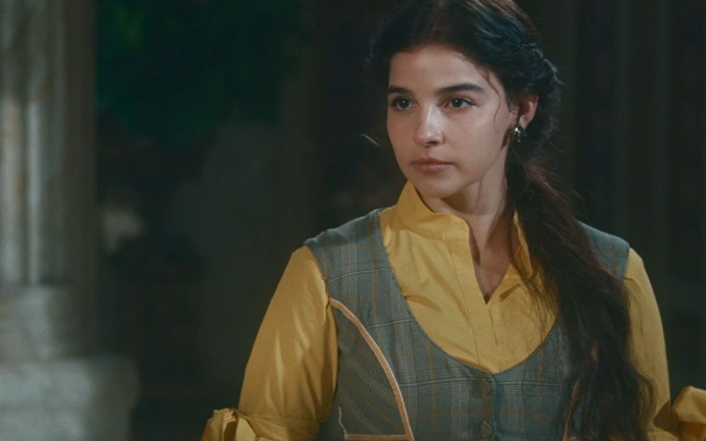 Gabriela Medvedovski grava cena com vestido em tons de amarelo e cinza, cabelo preso e expressão séria, como Pilar