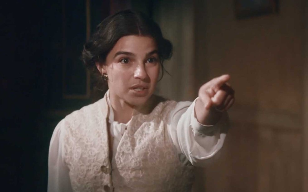 A atriz Gabriela Medvedovski com expressão de desespero aponta com o dedo indicador da mão direita para a direita em cena de Nos Tempos do imperador