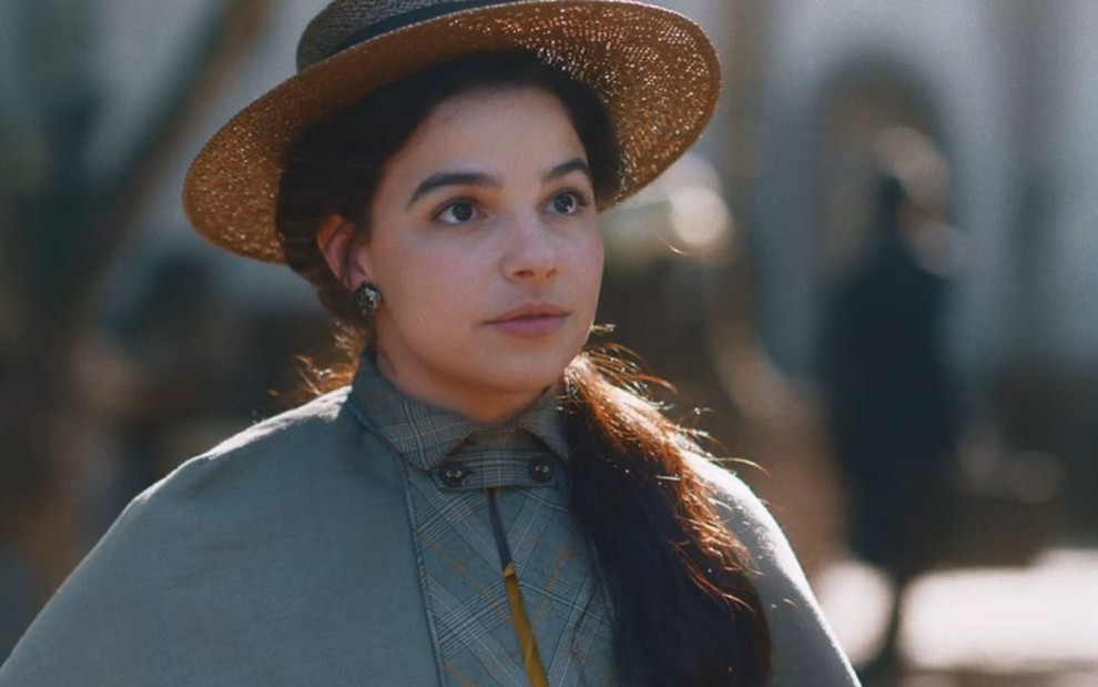 Pilar (Gabriela Medvedovski) usa chapéu; ela está parada em rua em cena de Nos Tempos do Imperador