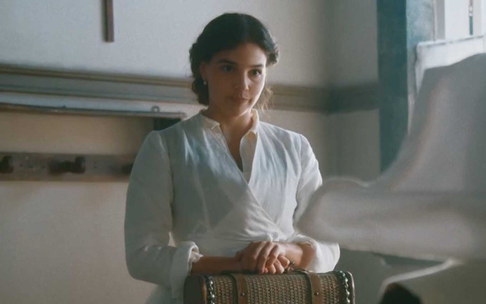 A atriz Gabriela Medvedovski com um vestido branco, segura uma maleta de médica, como a Pilar em cena de Nos Tempos do Imperador