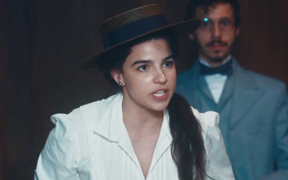 A atriz Gabriela Medvedovski como Pilar tem uma expressão de raiva com João Pedro Zappa, o Nélio, ao fundo em cena de Nos Tempos do Imperador
