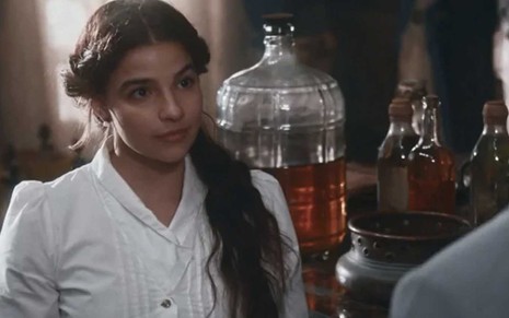 A atriz Gabriela Medvedovski como Pilar vestida de médica e em frente a vários fracos de remédio emc ena