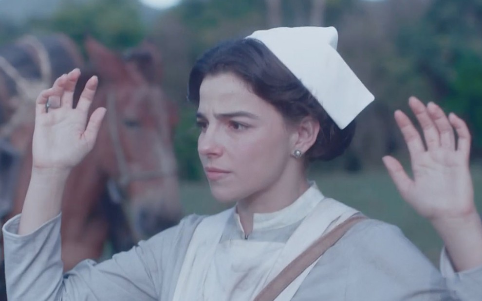 A atriz Gabriela Medvedovski com uniforme de enfermeira, na pele de Pilar, levanta as mãos para cima ao ser rendida em cena de Nos Tempos do Imperador