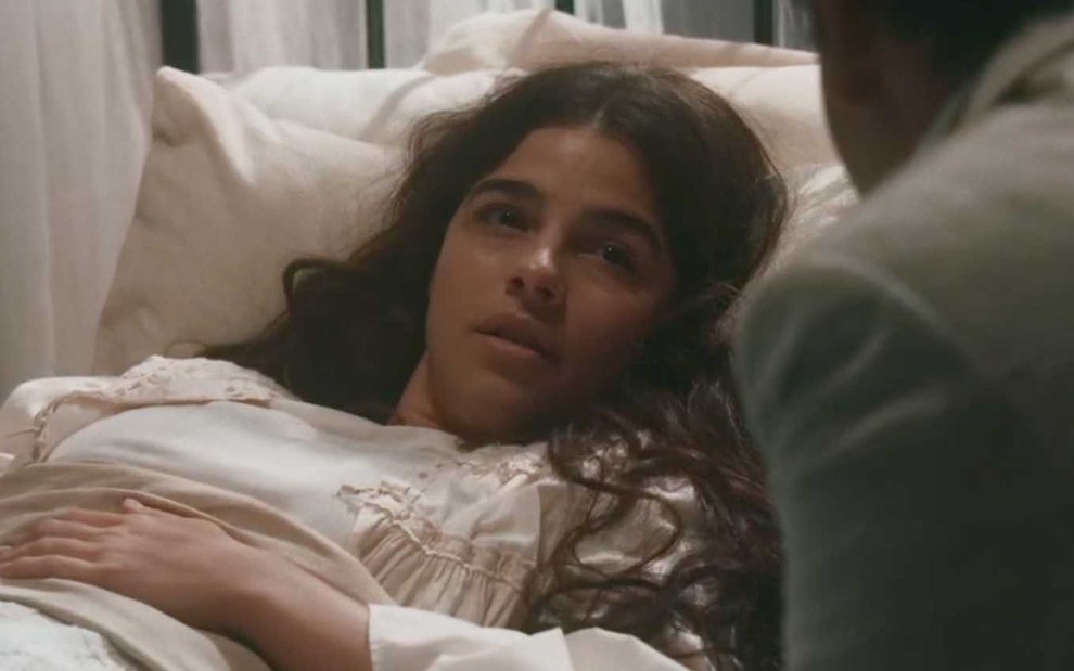 A atriz Gabriela Medvedovski caracterizada como Pilar deitada em uma cama com expressão de fragilidade em cena de Nos Tempos do Imperador
