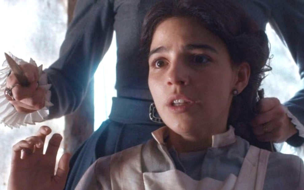 Pilar (Gabriela Medvedovski) está ajoelhada, e a mão de Elisa (Lana Rhodes) com uma faca aparece atrás dela em cena de Nos Tempos do Imperador