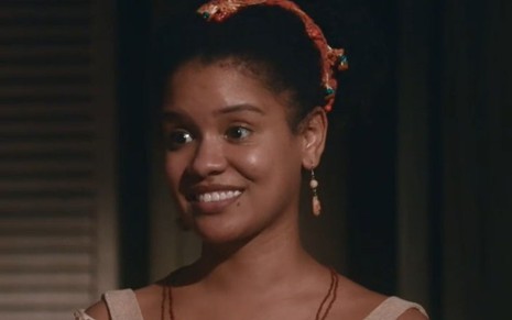 Zayla (Heslaine Vieira) sorri e está em pé na sala de cassino em cena de Nos Tempos do Imperador