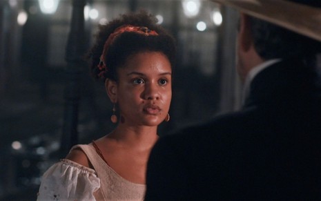 Heslaine Vieira está caracterizada como Zayla e conversa com o intérprete de Tonico, Alexandre Nero, que usa chapéu e está de costas para a foto em cena de Nos Tempos do Imperador