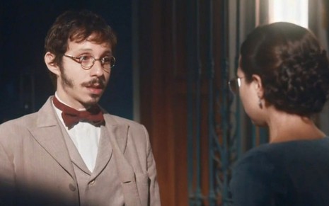 Nélio (João Pedro Zappa) está em pé na sala de Dolores (Daphne Bozaski), que está de costas na foto; os dois conversam