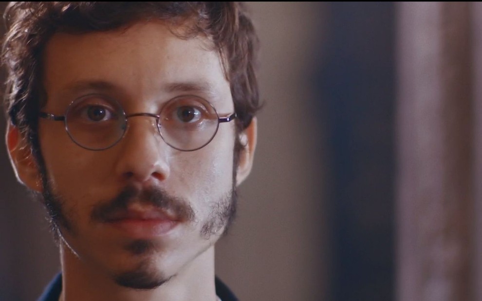 Nélio (João Pedro Zappa) usa óculos e está parado em sala em cena de Nos Tempos do Imperador