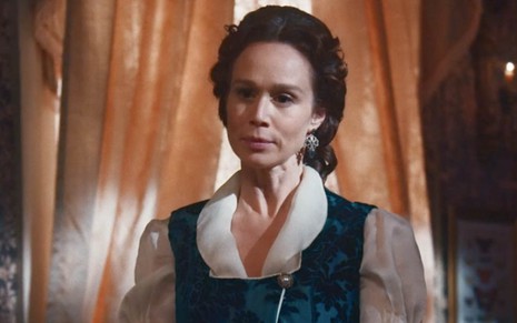 A atriz Mariana Ximenes está em uma sala, com a janela tapada parcialmente por cortinas ao fundo,  caracterizada como a Luísa em cena de Nos Tempos do Imperador