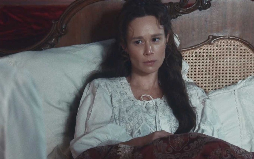 A atriz Mariana Ximenes como Luísa, sentada em uma cama, com aspecto de adoentada em cena de Nos Tempos do Imperador