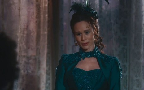 Luísa (Mariana Ximenes) está em pé na sala de sua casa; ela usa um vestido verde e um enfeite no cabelo em cena de Nos Tempos do Imperador