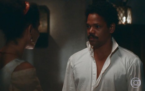 Samuel (Michel Gomes) está em pé na sala da sua casa com Zayla (Heslaine Vieira), que está de costas na foto, em cena de Nos Tempos do Imperador