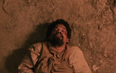 O ator Michel Gomes caracterizado como Samuel está machucado e caído no chão de barro em cena de Nos Tempos do Imperador