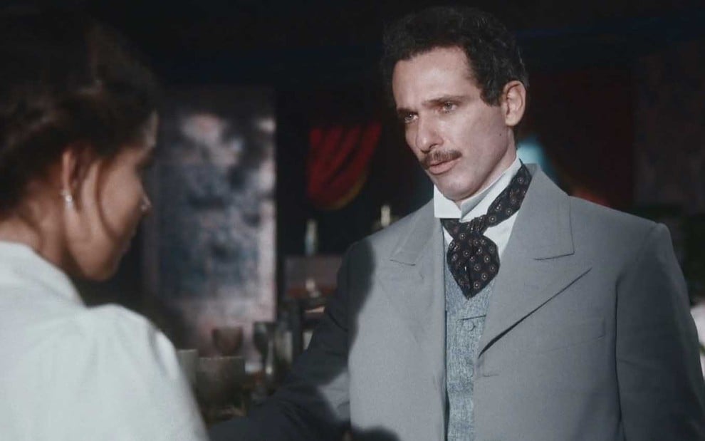 O ator Mouhamed Harfouch com um terno cinza e uma gravata azul de bolinhas como o Diego em cena de Nos Tempos do Imperador