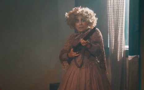 A atriz Paula Cohen como Lota, com uma peruca loira, cacheada e desgrenhada, segura uma espingarda com as duas mãos e com cara de assustada em cena de Nos Tempos do Imperador