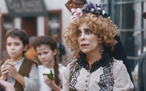 A atriz Paula Cohen com maquiagem exagerada e uma peruca cacheada loira, volumosa, com expressão de tristeza como a Lota em cena de Nos Tempos do Imperador