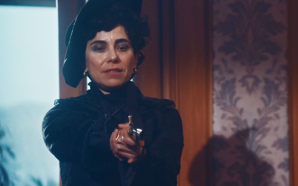 A atriz Paula Cohen, vestida de preto em luto, como a Lota, aponta uma arma