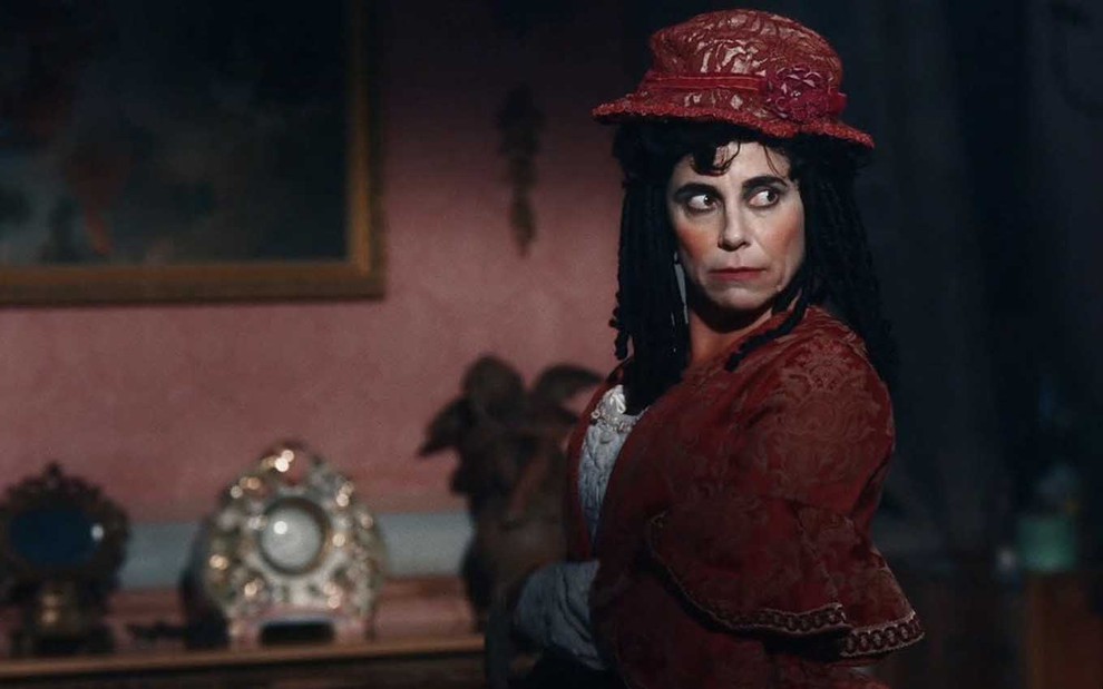 A atriz Paula Cohen com maquiagem exagerada, vestido de renda vermelho, igualmente exagerado, e uma peruca preta e cacheada como a Lota em cena de Nos Tempos do Imperador