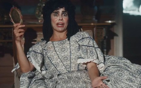 Lota (Paula Cohen) está sentada em sofá e segura um espelho em cena de Nos Tempos do Imperador
