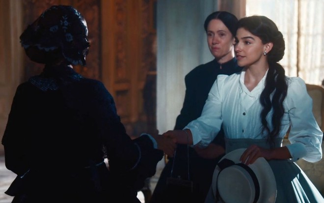 Gabriela Medvedovski caracterizada como Pilar em cena de Nos Tempos do Imperador: atriz está com vestido branco e aperta mão de personagem de costas