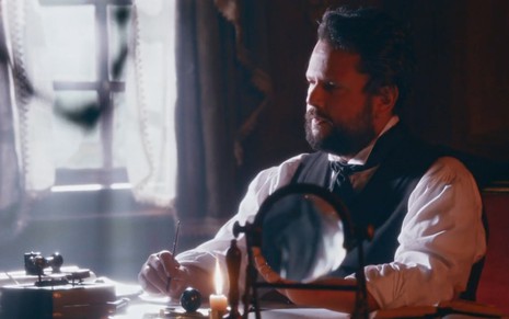 O ator Selton Mello como Pedro sentado à mesa escrevendo uma carta em um escritório em cena vespertina de Nos Tempos do Imperador
