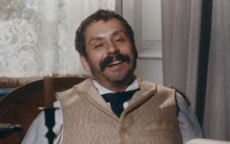 Alexandre Nero em cena de Nos Tempos do Imperador: ator está sentado, caracterizado como Tonico e sorri
