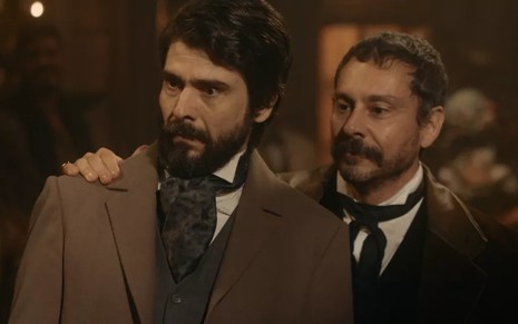 Lucci Ferreira, de terno marrom e expressão de pavor, grava ao lado de Alexandre Nero, com sorriso, como Floriano e Tonico