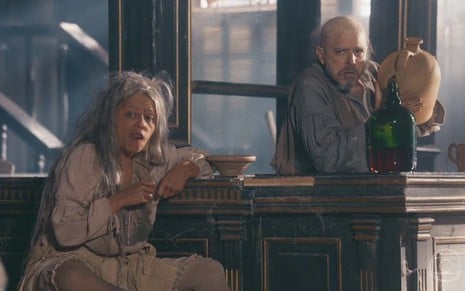 A atriz Vivianne Pasmanter como Germana está sentada em um banco em frente a um balcão enquanto Guilherme Piva, o Licurgo, vira um galã de vinho em uma garrafa de vidro em cena de Nos Tempos do Imperador