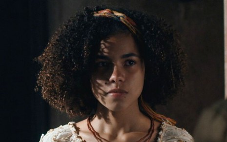 Alana Cabral em cena de Nos Tempos do Imperador: atriz está caracterizada como Zayla, com um vestido branco e colar marrom, e olha com desconfiança