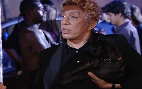 Clodovil Hernandes usa camisa e blazer pretos; com um pug preto no colo, ele encara Alicinha (Cristiana Oliveira) --fora do quadro-- com deboche em cena de O Clone