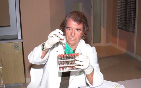 O ator Juca de Oliveira caracterizado como Albieri mexe em tubos de ensaio com as mãos protegidas por luvas cirúrgicas brancas em cena de O Clone