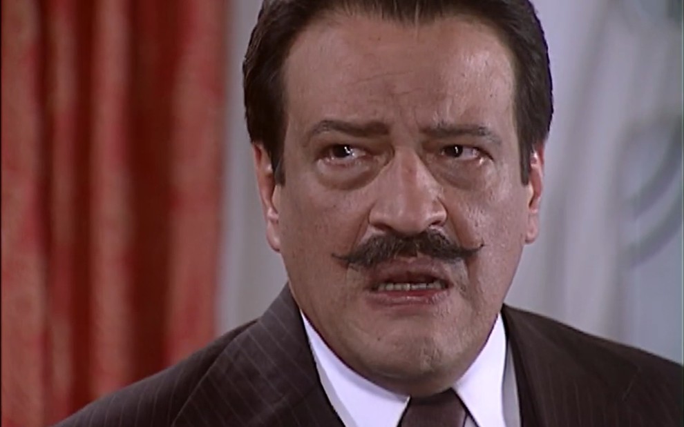 Luís Melo, caracterizado como Batista, olha para o lado, com as sobrancelhas franzidas e a expressão irritada em cena de O Cravo e a Rosa