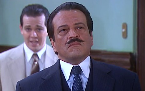 Luís Melo, caracterizado como Batista, tem a expressão pensativa; ao fundo, Teodoro (Matheus Petinatti) se justifica em cena de O Cravo e a Rosa