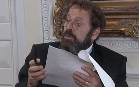 Carlos Vereza, caracterizado como Joaquim, segura um papel com a expressão debochada em cena de O Cravo e a Rosa
