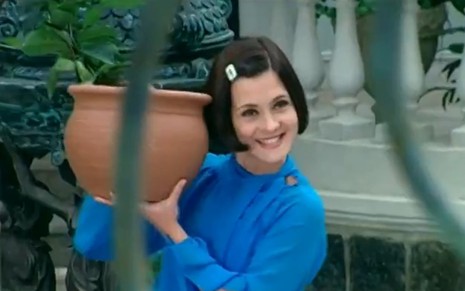 Adriana Esteves, caracterizada como Catarina, sorri enquanto segura um vaso em cena de O Cravo e a Rosa