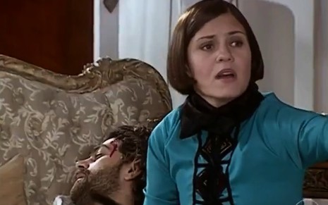 Adriana Esteves, caracterizada como Catarina, olha para além da câmera, desesperada; ao fundo, é possível ver Eduardo Moscovis, o Petruchio, com um ferimento na cabeça