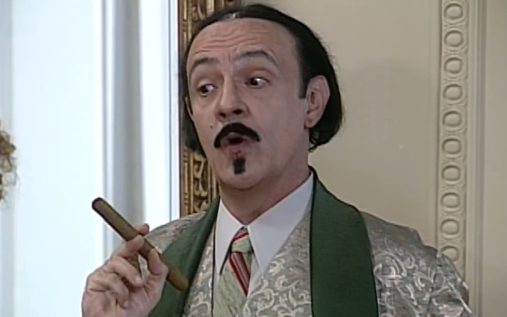 Ney Latorraca caracterizado como Cornélio: ator tem cabelo na altura dos queixo e usa bigode e cavanhaque. Ele tem a expressão debochada enquanto segura um charuto em cena de O Cravo e a Rosa