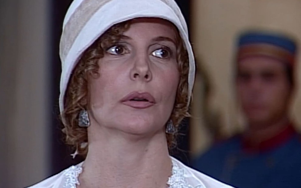 Maria Padilha tem os cabelos na altura do queixo, cobertos com um chapéu branco. Ela olha para o lado, com a boca apertada. A maquiagem é leve