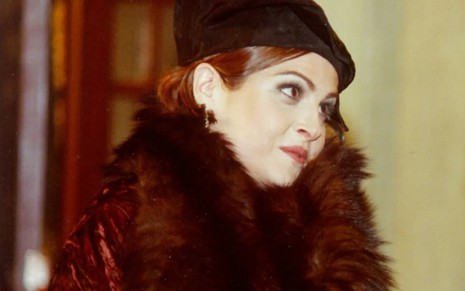 Drica Moraes tem os cabelos curtos, ruivos, cobertos por um chapéu de veludo. Ela veste um enorme casaco de pelos preto. De perfil, a atriz dá um leve sorriso e olha para o lado durante cena de O Cravo e a Rosa