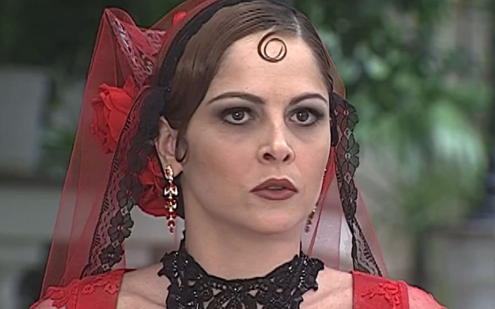 Drica Moraes, caracterizada como Marcela, tem a expressão raivosa em cena de O Cravo e a Rosa; ela usa um vestido de noiva vermelho e um grande colar preto. A maquiagem é forte, com olhos bem marcantes e um batom vermelho.