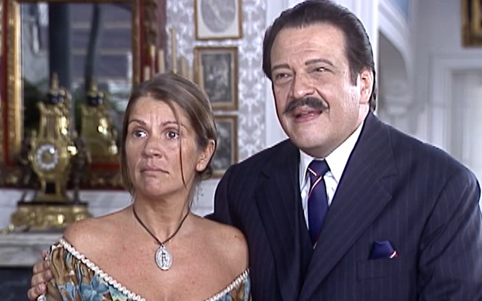 Tassia Camargo, caracterizada como Joana, tem a expressão chocada;  Luís Melo, o Batista, sorri em cena de O Cravo e a Rosa