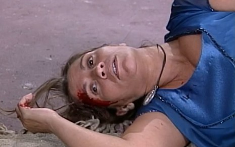 Tássia Camargo está estirada no chão; ela está pálida, com o lado direito da cabeça cheio de sangue, em cena de O Cravo e a Rosa