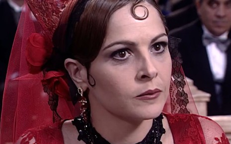Drica Moraes caracterizada como Marcela: atriz usa um vestido de noiva vermelho cheio de renda. Ela ainda aderiu a brincos e colar preto. Um baby hair forma um cacho moldado na testa da atriz em cena de O Cravo e a Rosa