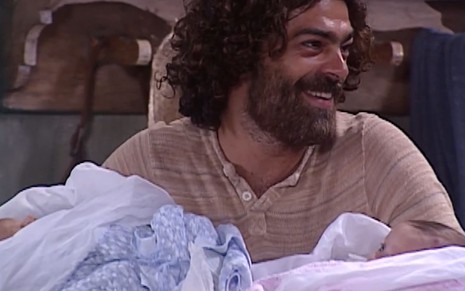 Eduardo Moscovis caracterizado como Petruchio: cabelo e barba longos, sobrancelha por fazer e blusa surrada. Ele dá um sorriso largo e segura dois bebês no colo --um, enrolado em panos azuis; outro, em um cobertor rosa.
